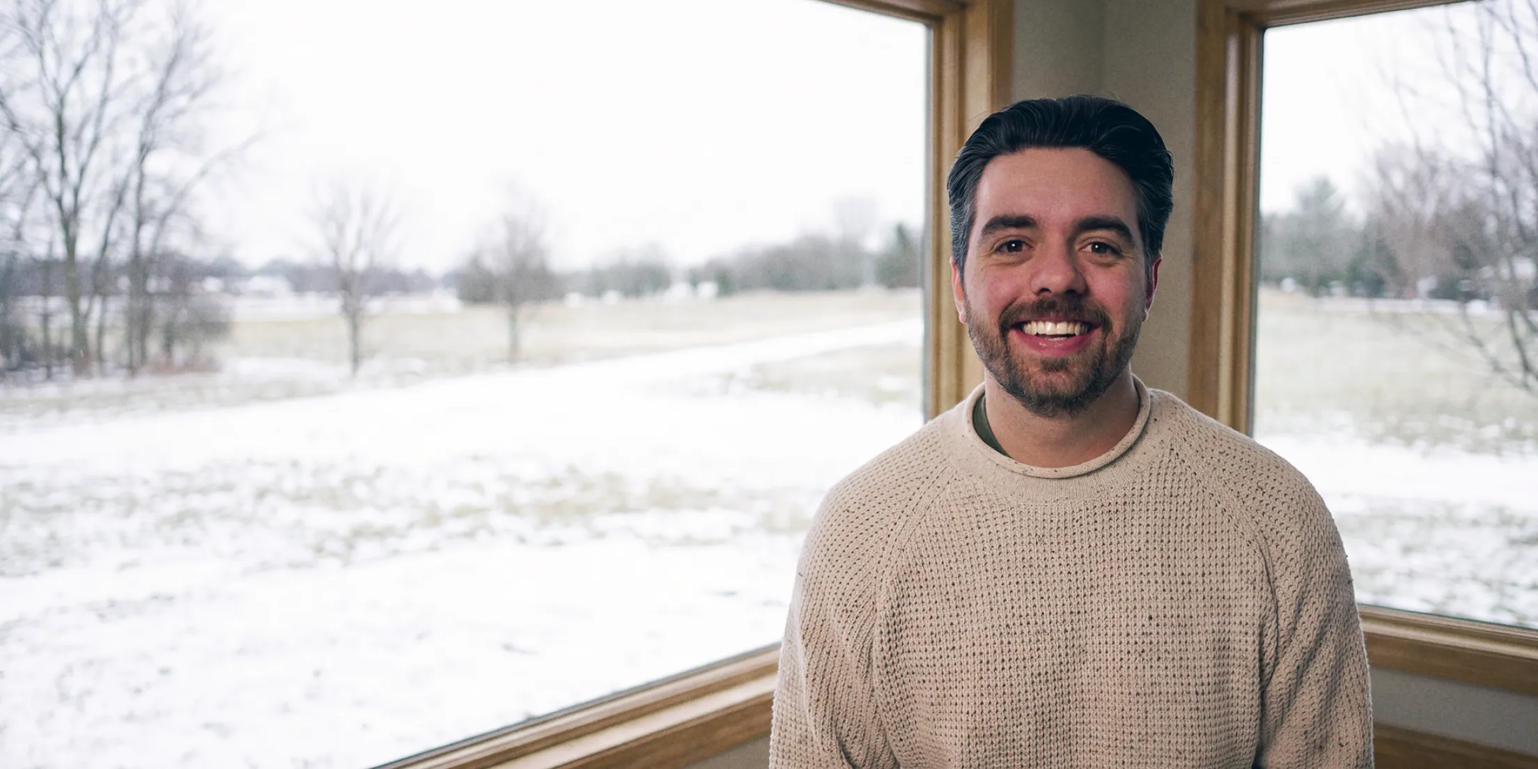 Kyle Sundman in a knit sweater, smiling in a corner room with windows onto a snowy field